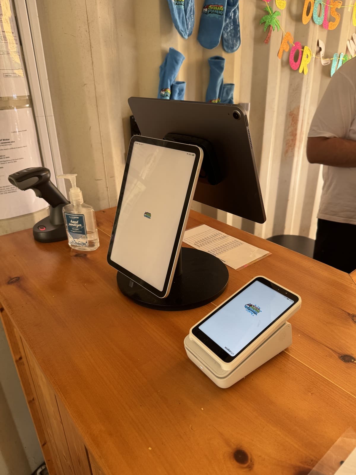 A cash register set up with the Bouncepad dual stand (customer-facing iPad in front, staff iPad behind), a card payment terminal, and a barcode scanner