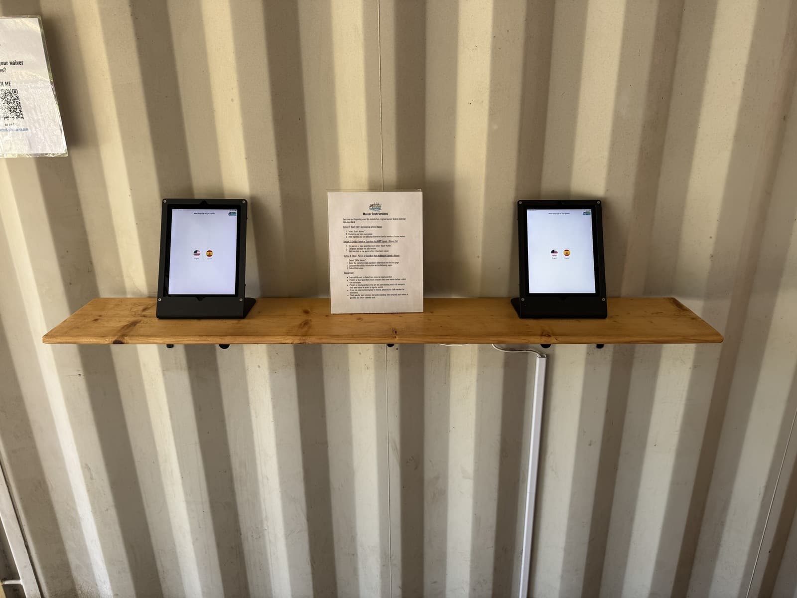 A waiver station: two single Bouncepad tablet stands on a shelf, each showing the language selection, with a printed instructions sheet between them